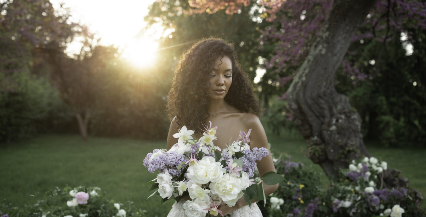 Bride with voluminous lilac and peony bouquet at golden hour in a château garden, wearing a lace bustier gown with loose curls.