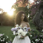 Bride with voluminous lilac and peony bouquet at golden hour in a château garden, wearing a lace bustier gown with loose curls.