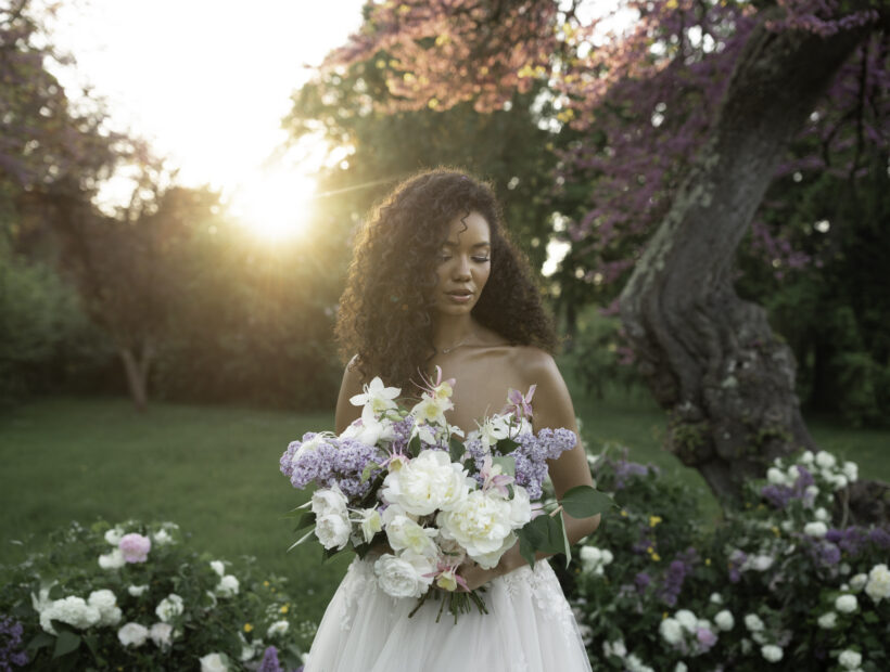 Bride with voluminous lilac and peony bouquet at golden hour in a château garden, wearing a lace bustier gown with loose curls.