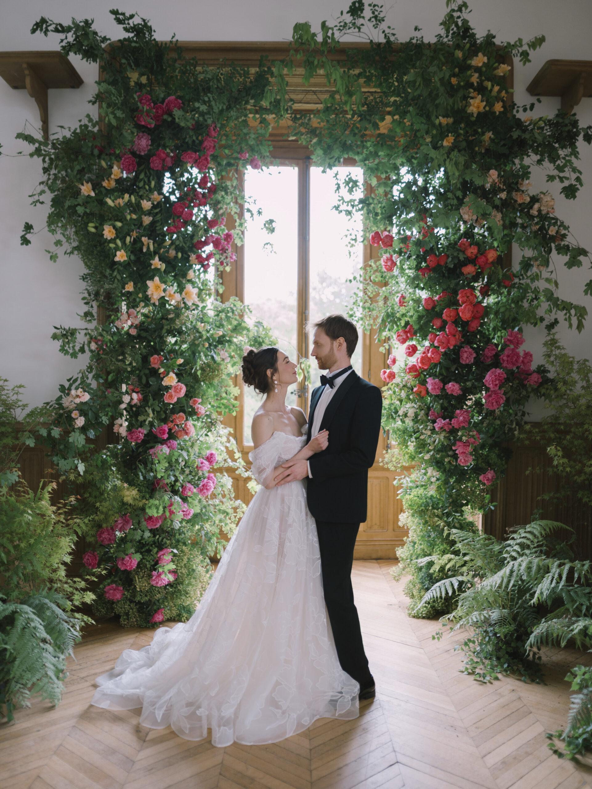 Bride and groom embracing beneath a lush floral installation filled with roses and greenery at Château de Méridon, France. Elegant garden-inspired wedding design for a luxury destination wedding near Paris.