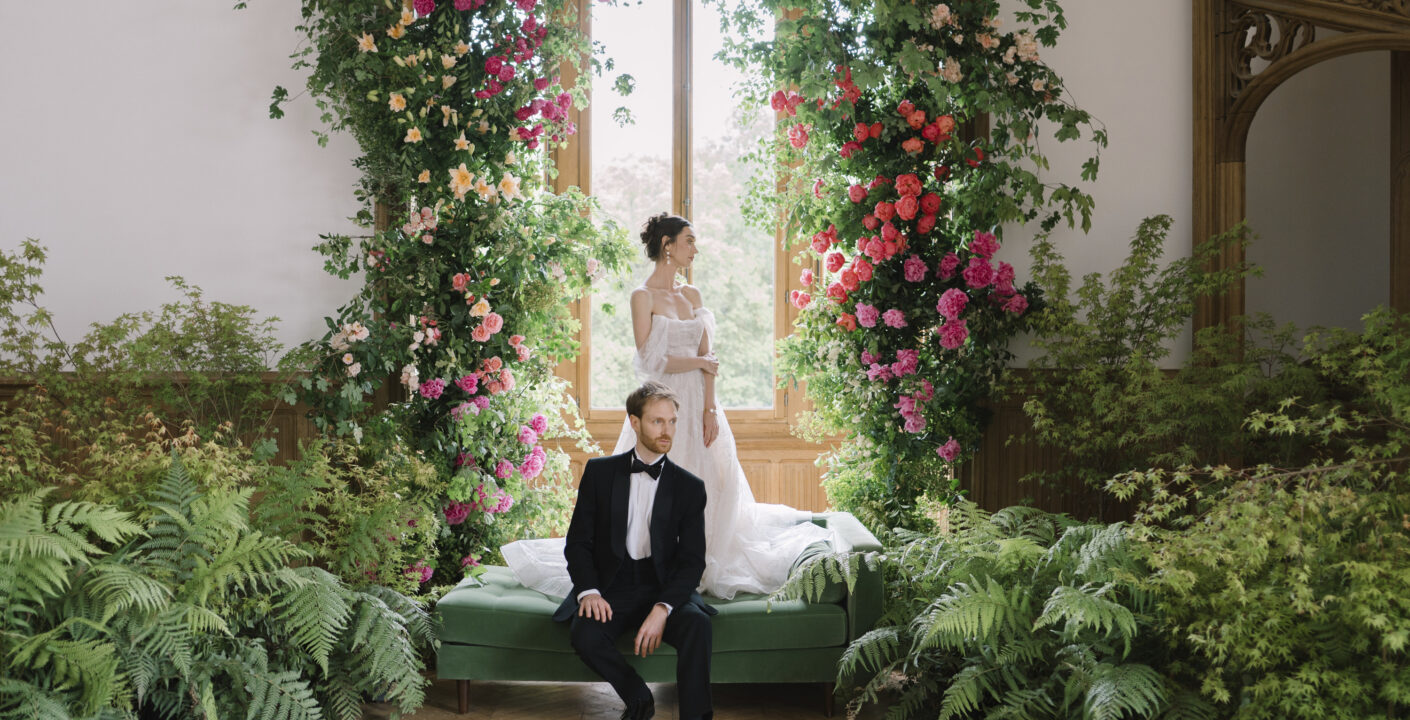 Bride and groom posing in an elegant French château salon framed by a lush floral installation of climbing roses, greenery, and ferns, creating a romantic garden-inspired wedding.