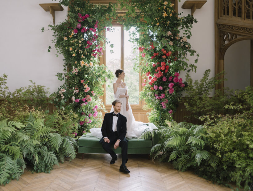 Bride and groom posing in an elegant French château salon framed by a lush floral installation of climbing roses, greenery, and ferns, creating a romantic garden-inspired wedding.