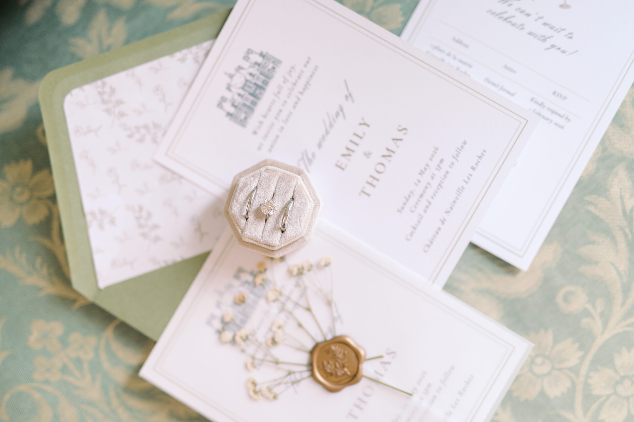 Elegant wedding invitation suite for Emily and Thomas with a gold wax seal, baby's breath, and a diamond ring set in a velvet box, styled on a sage green damask backdrop. Wedding planning by Paris, I do.