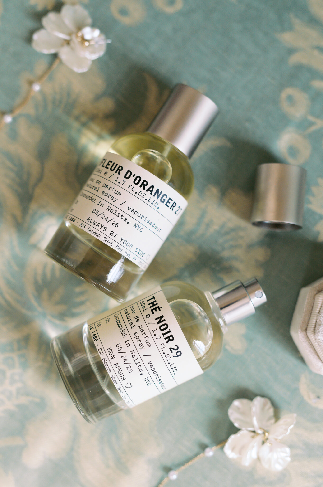 Two personalized wedding day perfume bottles with custom labels reading "Always by your side" and "Mon amour," styled with white floral details on a soft blue patterned fabric. Wedding planning by Paris, I do.