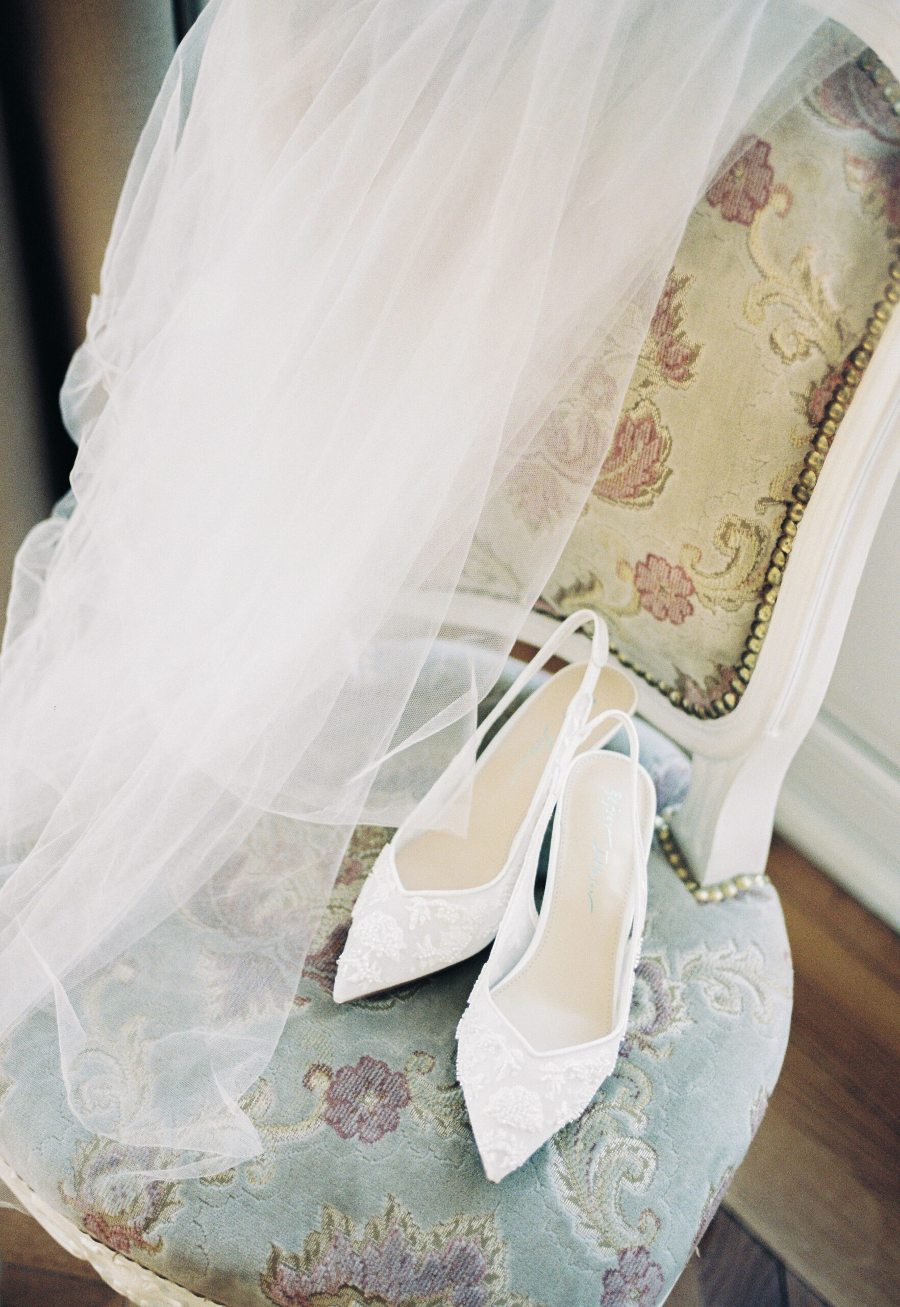 White lace-embellished bridal heels resting on a floral tapestry armchair, with a soft tulle veil draped beside them. Wedding planning by Paris, I do.