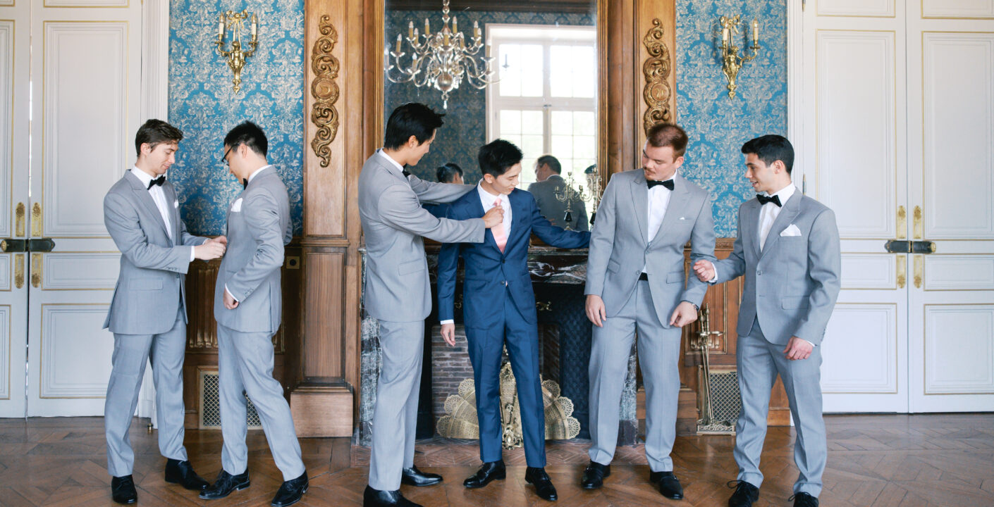 Groom in a navy blue suit with a blush tie, surrounded by groomsmen in light gray suits with black bow ties, getting ready in a château salon with blue damask walls and a gilded mirror. Wedding planning by Paris, I do.