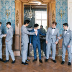 Groom in a navy blue suit with a blush tie, surrounded by groomsmen in light gray suits with black bow ties, getting ready in a château salon with blue damask walls and a gilded mirror. Wedding planning by Paris, I do.