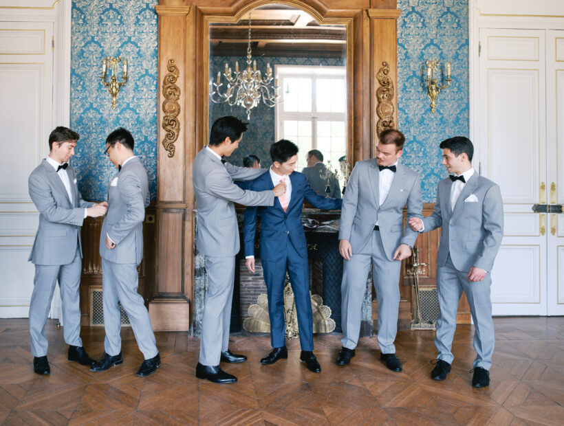 Groom in a navy blue suit with a blush tie, surrounded by groomsmen in light gray suits with black bow ties, getting ready in a château salon with blue damask walls and a gilded mirror. Wedding planning by Paris, I do.