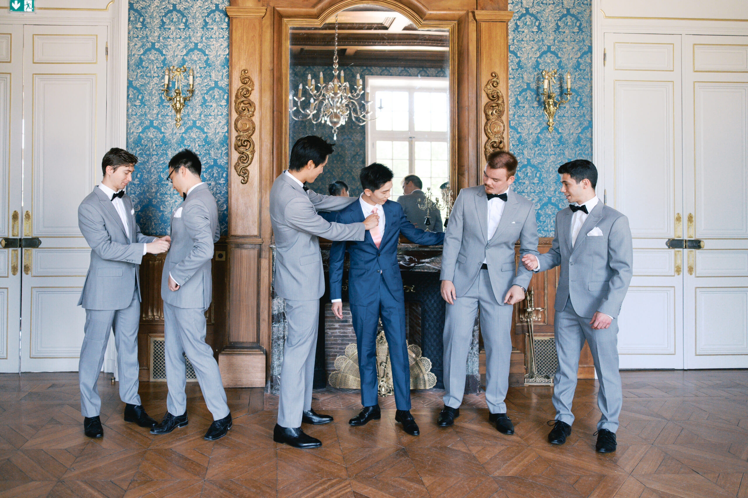Groom in a navy blue suit with a blush tie, surrounded by groomsmen in light gray suits with black bow ties, getting ready in a château salon with blue damask walls and a gilded mirror. Wedding planning by Paris, I do.