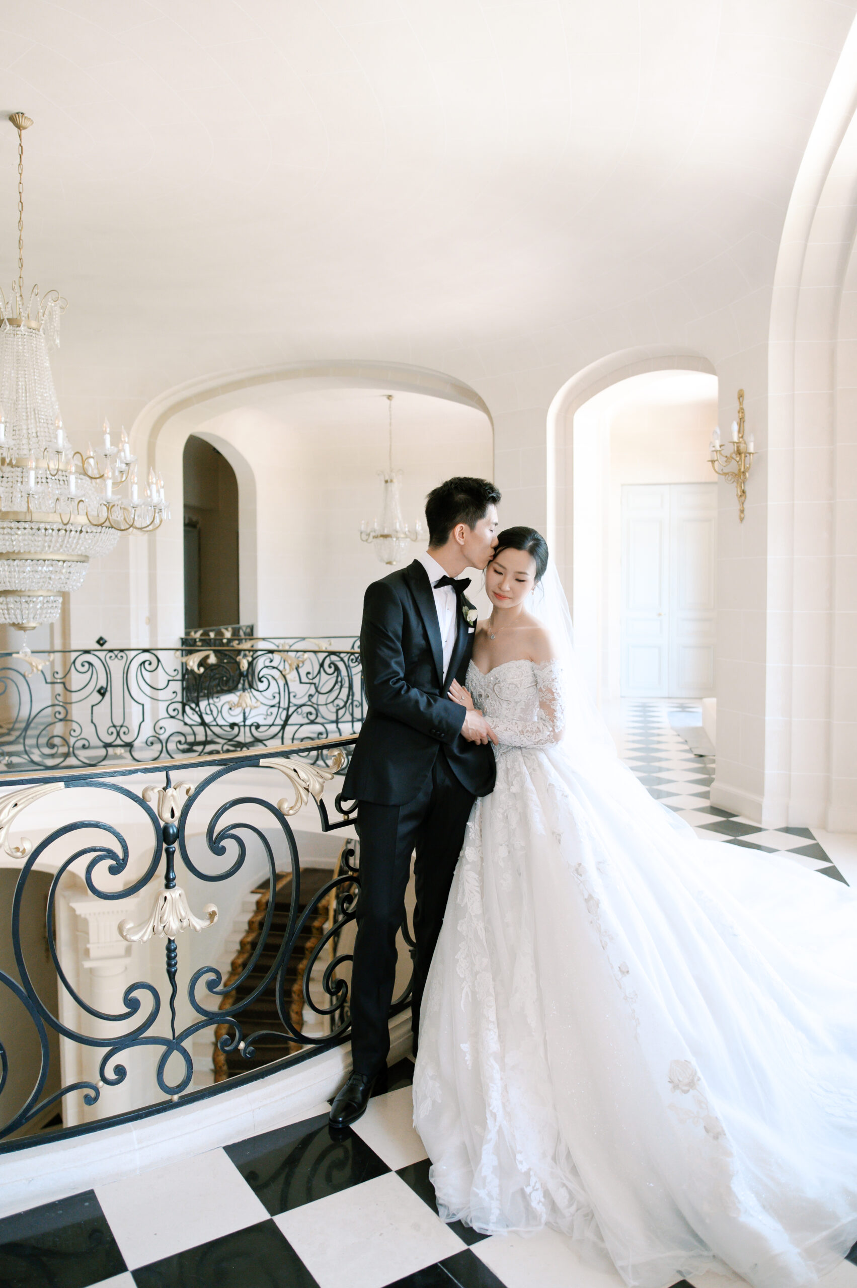 Groom in a black tuxedo kissing the forehead of his bride in a lace off-the-shoulder gown, on a grand château staircase with wrought-iron railing, crystal chandelier, and checkered marble floor. Wedding planning by Paris, I do.
